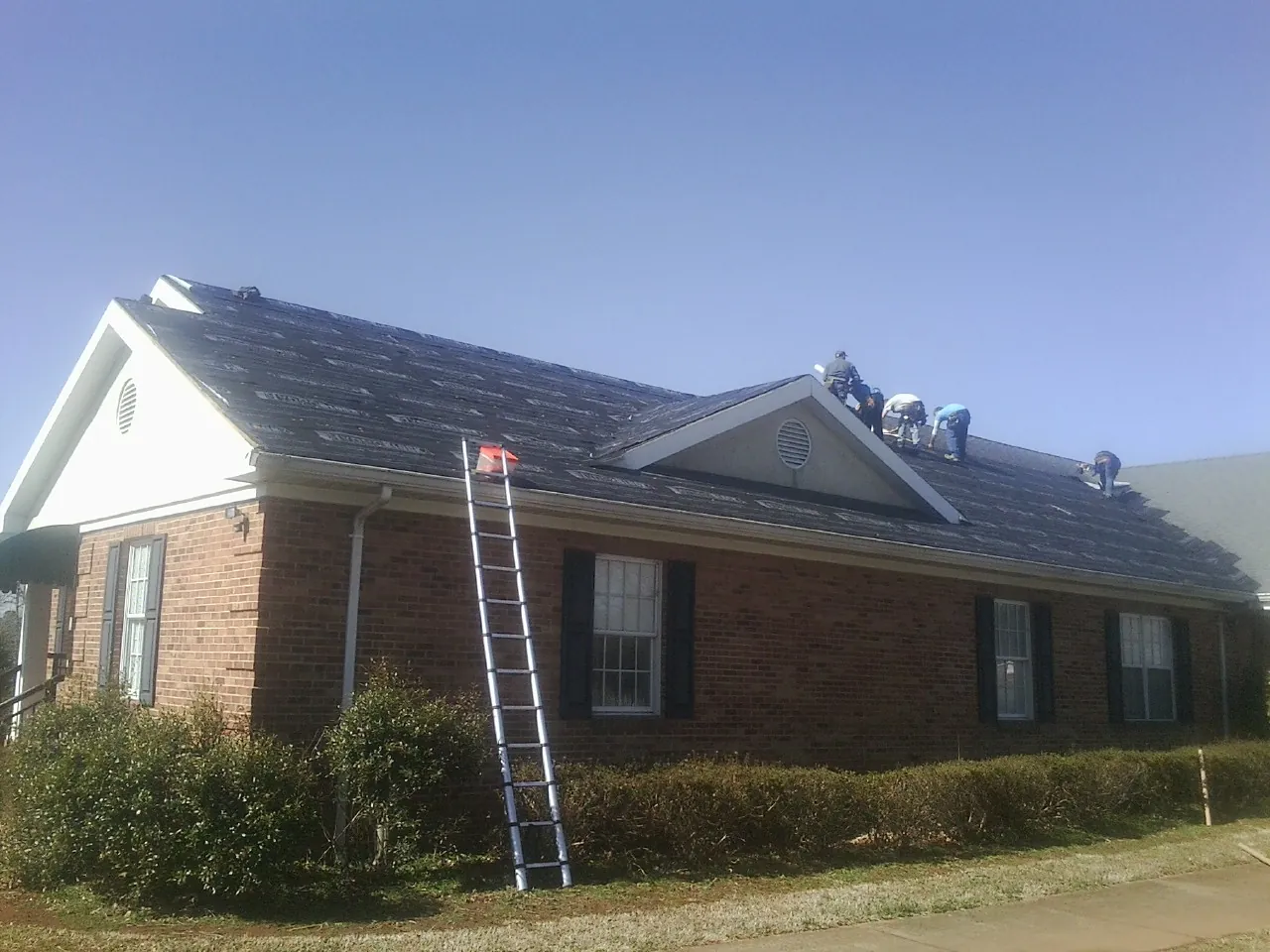 Crew tearing shingles off a brick ranch roof with a ladder set — Emergency Roof Repair work by our crew in Lamona
